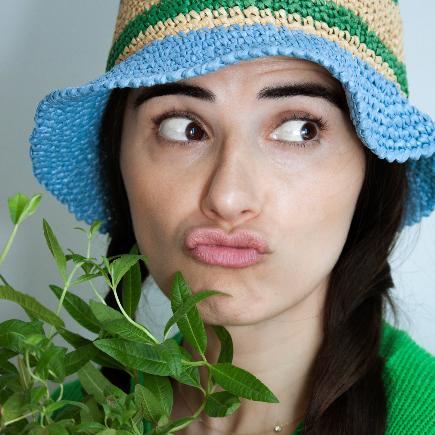 A woman wearing a colorful crochet hat makes a funny face while holding a fresh lemon verbena plant.