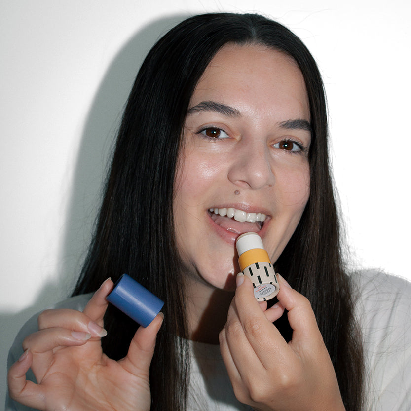Woman smiling while applying Zarbi vegan lip balm with a bold patterned tube and holding the blue cap, showcasing joyful clean beauty.