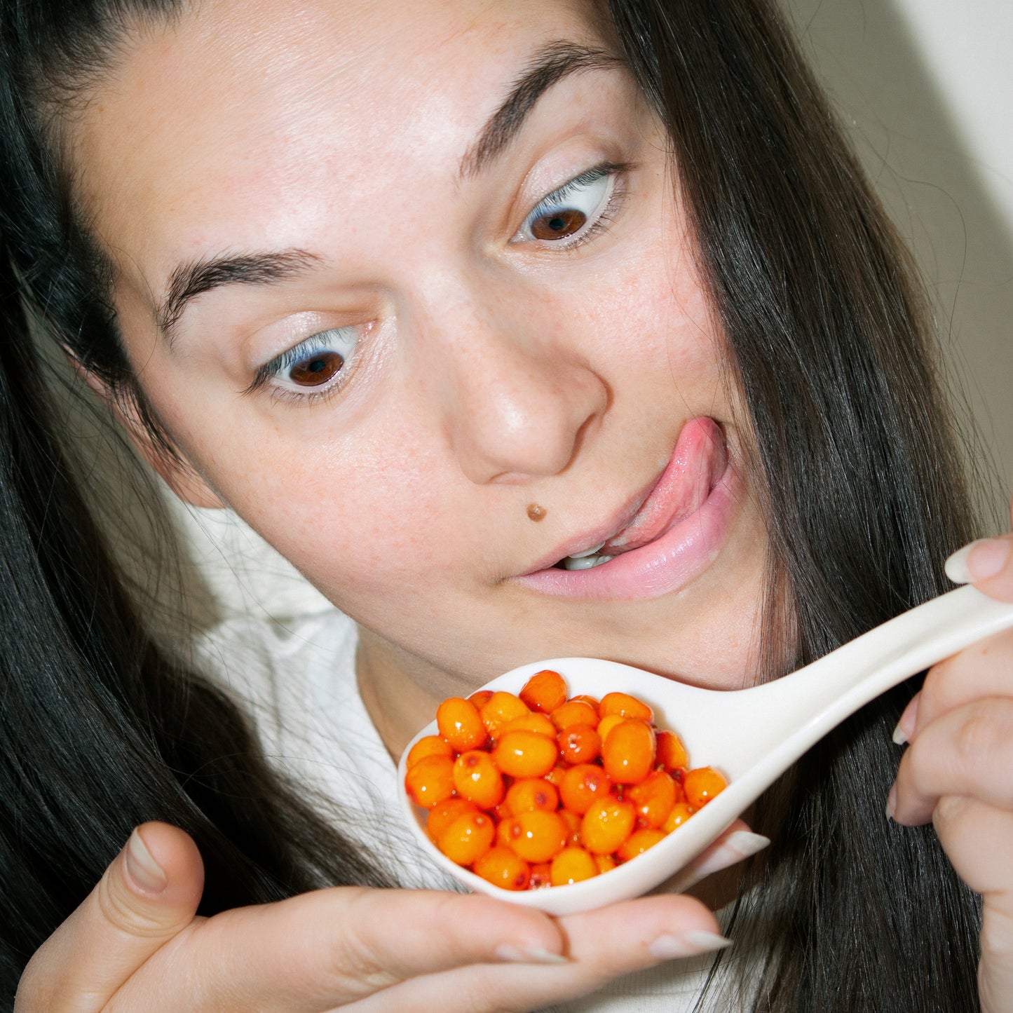 A woman makes a playful face while holding a spoonful of bright orange sea buckthorn berries close to her mouth.