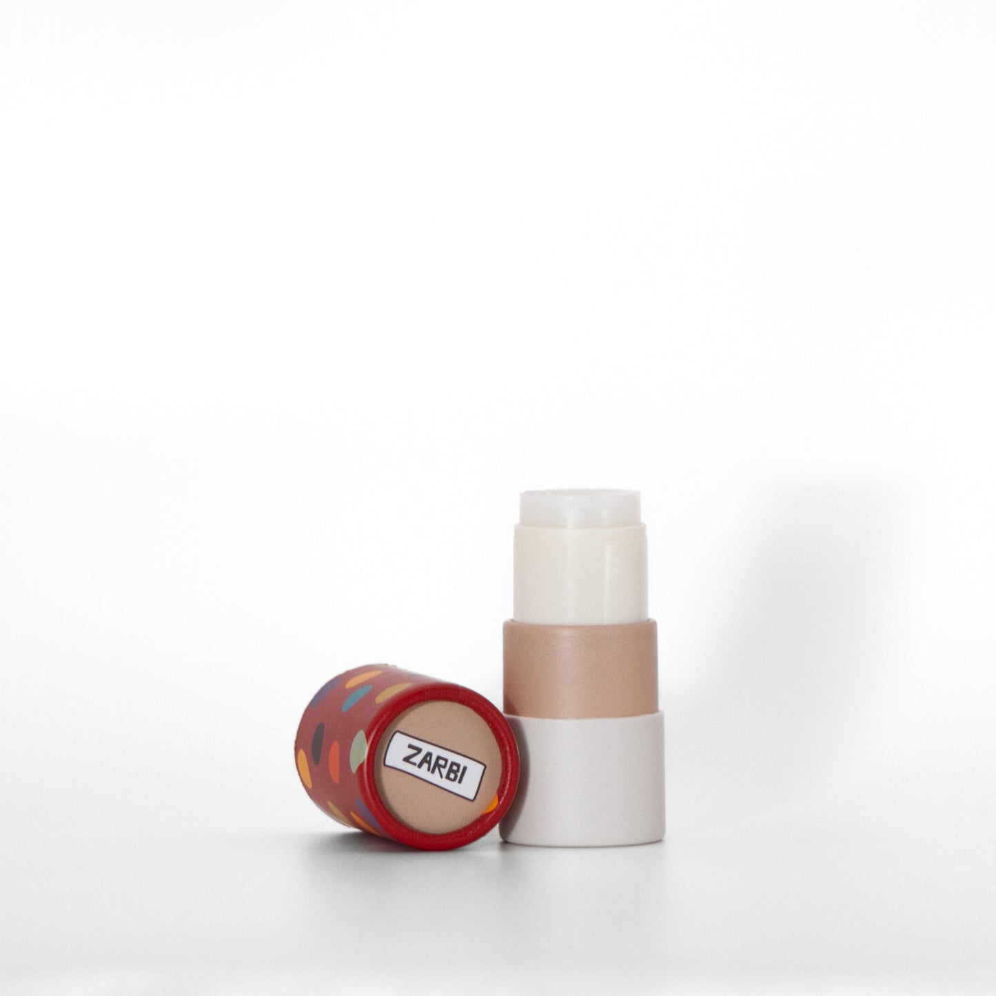 Open twist-up Banana Bubble Gum lip balm tube with cream base showing clear balm inside. Red cap with multicolor dots laying on the side with beige top showing Zarbi logo.