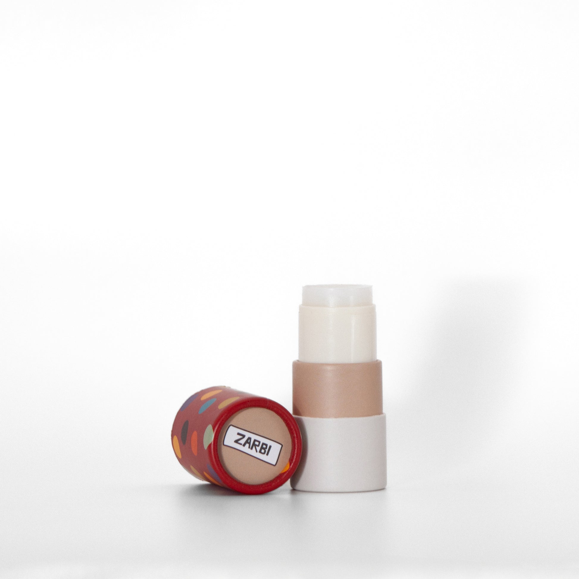 Open twist-up Banana Bubble Gum lip balm tube with cream base showing clear balm inside. Red cap with multicolor dots laying on the side with beige top showing Zarbi logo.