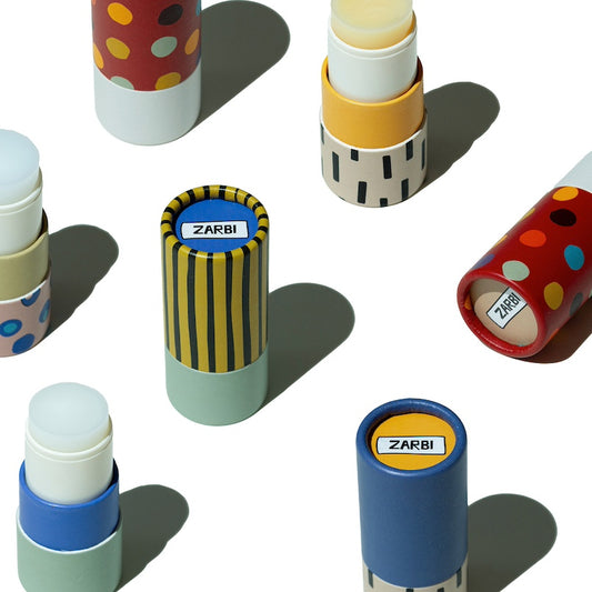 Several ZARBI lip balm tubes arranged on a white background, each showing different colorful packaging designs including red with yellow dots, yellow and black stripes, blue, beige, and red with multicolor dots. One tube is open, revealing a white balm stick.