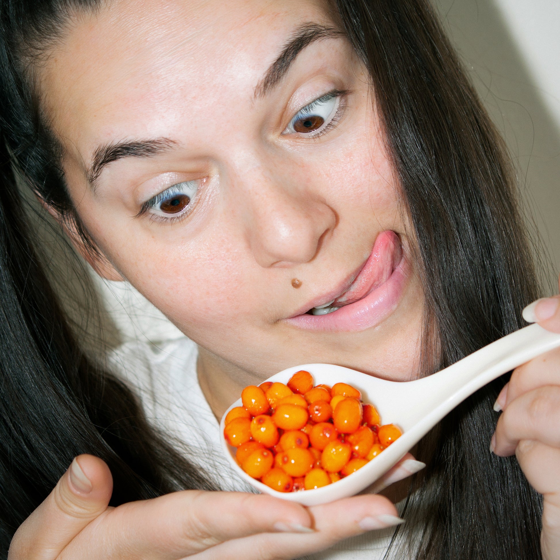 A woman makes a playful face while holding a spoonful of bright orange sea buckthorn berries close to her mouth.